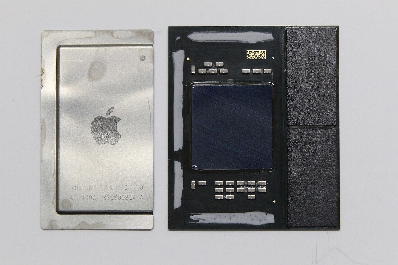 An Apple M1 with its metal lid removed and laid beside it: a dark square die in the middle of the package and two black memory packages along its right-hand edge