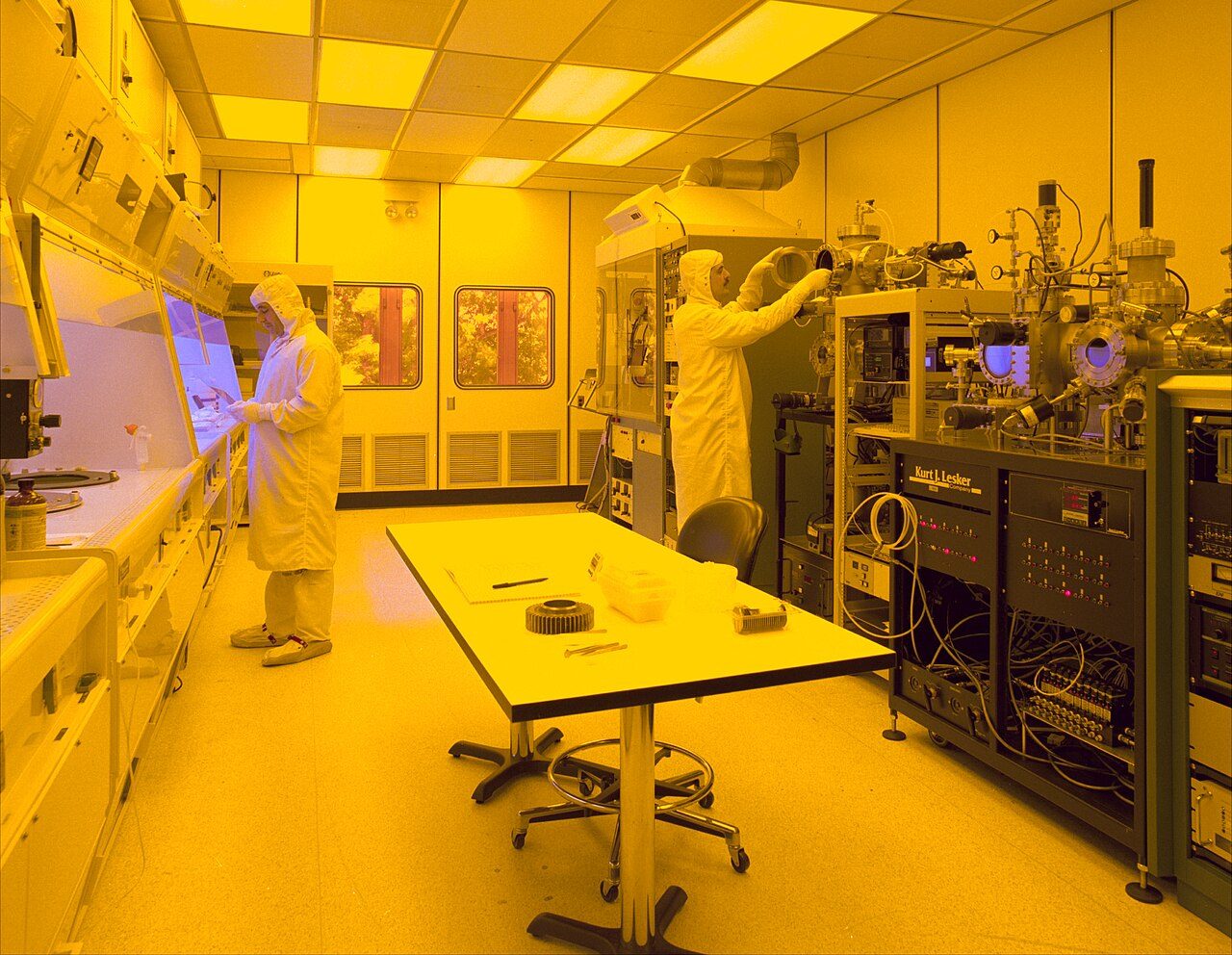 Two people in white cleanroom suits working at equipment in a room lit entirely in yellow