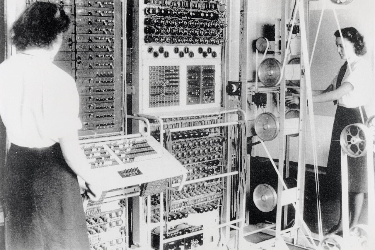 Two women operating Colossus: one at a panel of switches on the left, the other beside a tall frame of pulleys carrying a loop of paper tape on the right