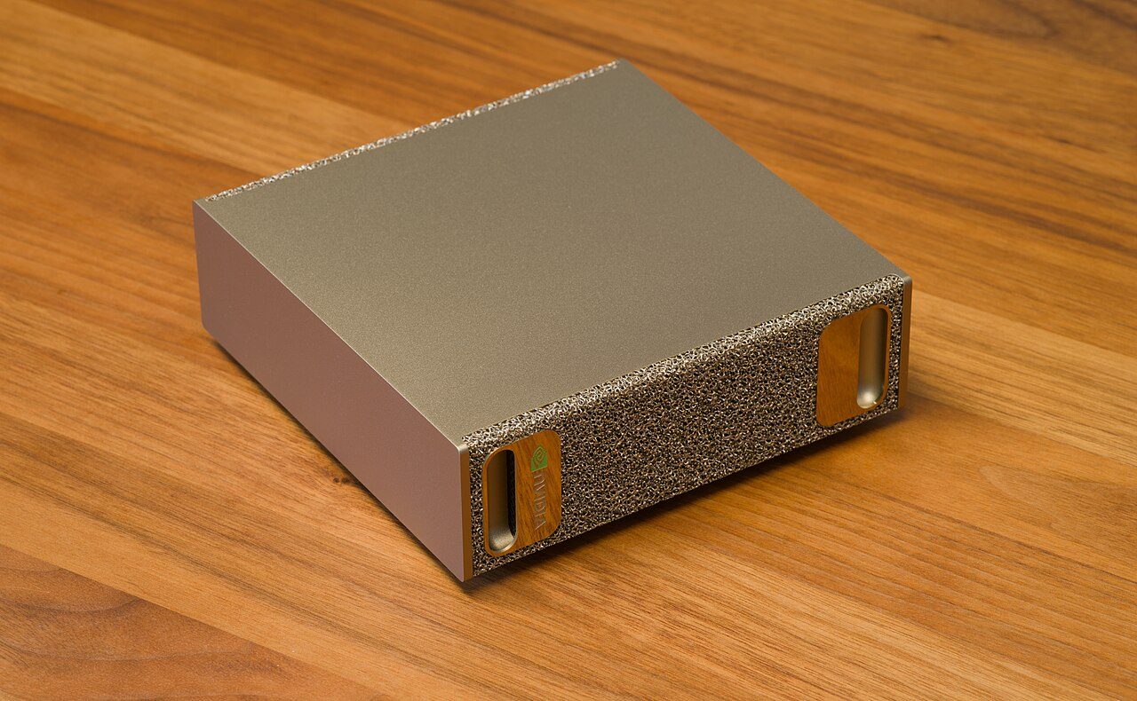 An Nvidia DGX Spark on a wooden desk: a small gold-coloured box about fifteen centimetres square with a metal-foam front panel