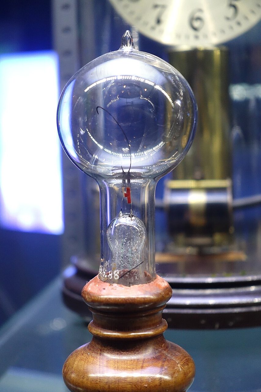A clear glass bulb with a thin looped filament inside, on a turned wooden base, in a museum case