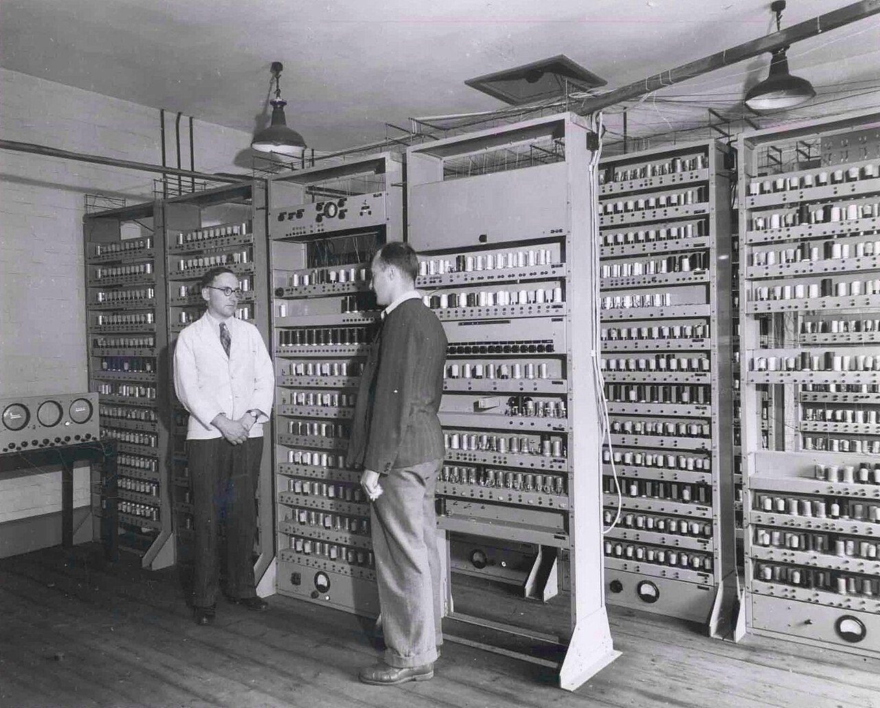 Two men standing in front of EDSAC, a room of tall open racks filled with vacuum tubes