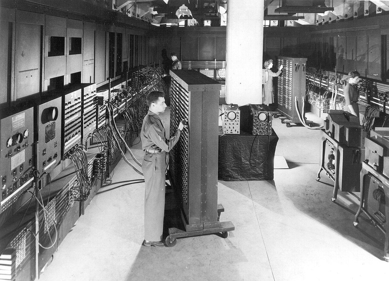ENIAC at the Moore School: a room lined with tall panels of switches, cables and dials, with a soldier setting switches on a wheeled function table in the foreground