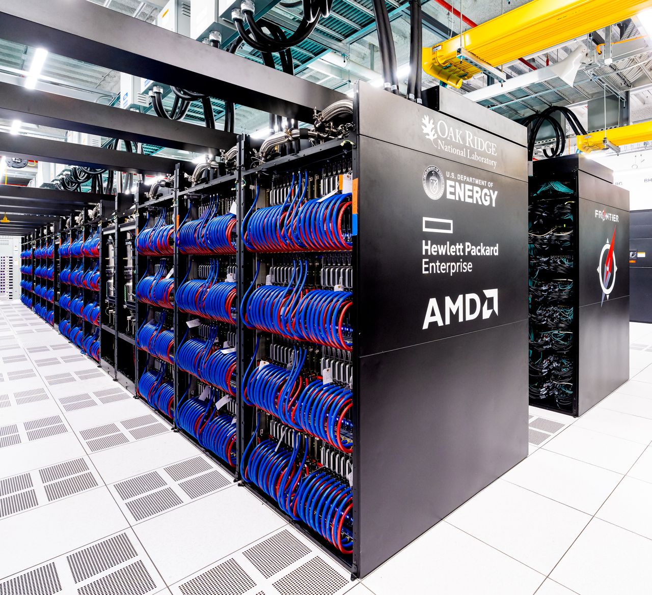 Rows of black Frontier supercomputer cabinets in a white machine room, the nearest end panel showing the Frontier logo with Oak Ridge, US Department of Energy, Hewlett Packard Enterprise and AMD marks, and bundles of blue cables visible through the open sides