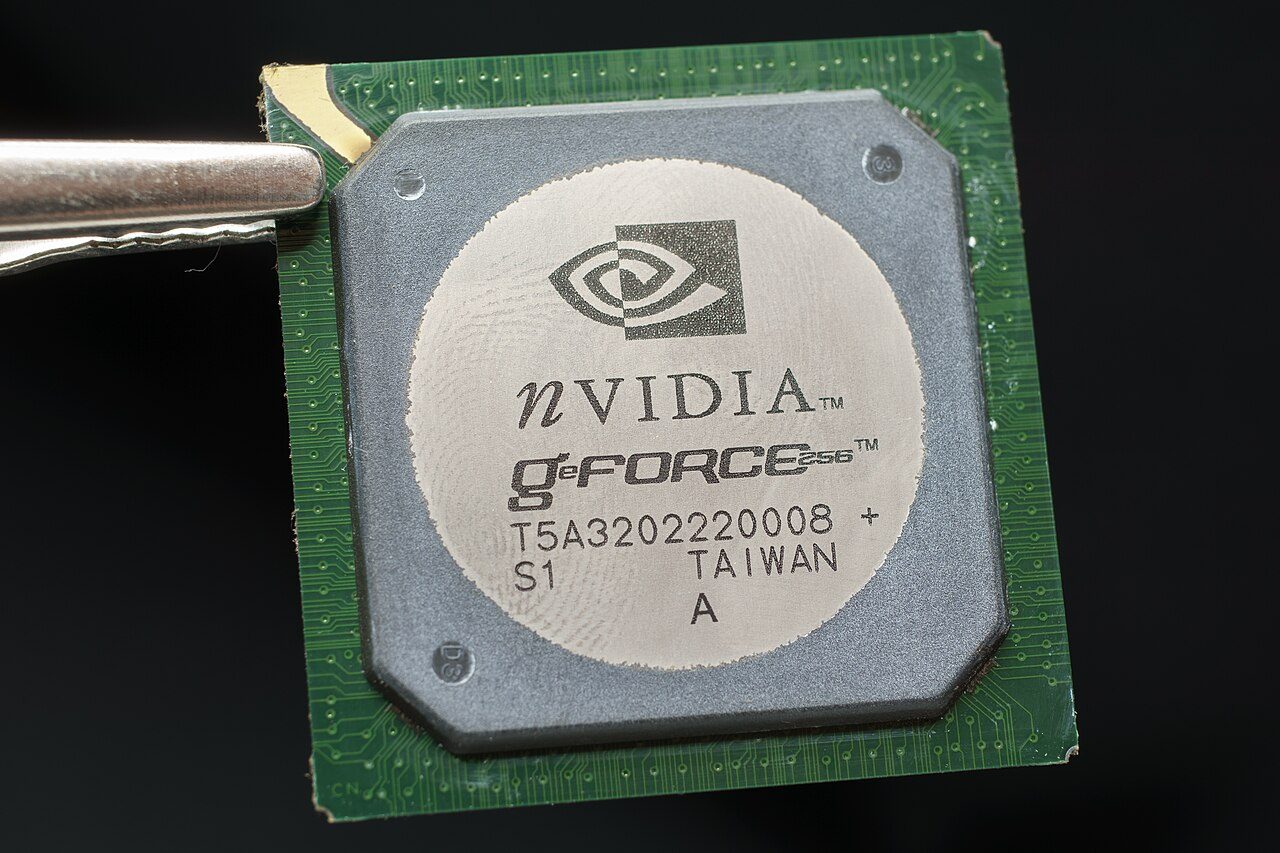 A GeForce 256 chip held in tweezers: a square green package with a round silver heat spreader printed with the Nvidia logo and GeForce 256
