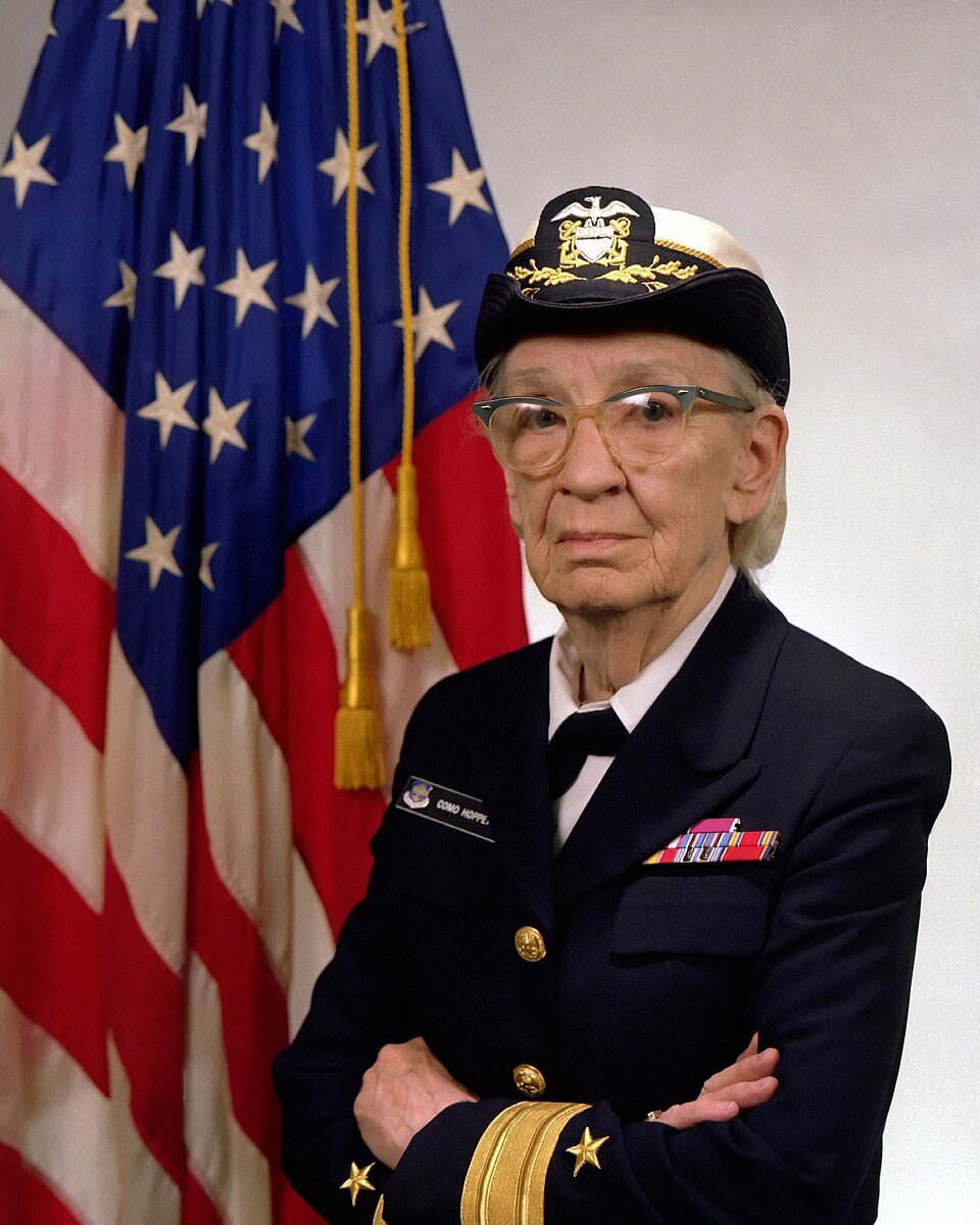Official portrait of Grace Hopper in US Navy uniform and cap in front of an American flag