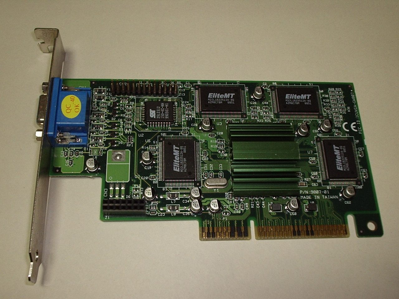 A green Intel740 graphics card for the AGP slot on a beige background, with a blue VGA connector on its metal bracket, four black memory chips marked EliteMT, and a finned heatsink in the middle over the graphics chip
