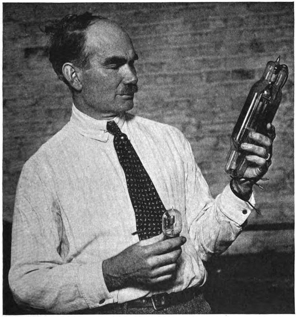 Lee de Forest in shirtsleeves and tie holding a small receiving tube in one hand and a large transmitting tube in the other