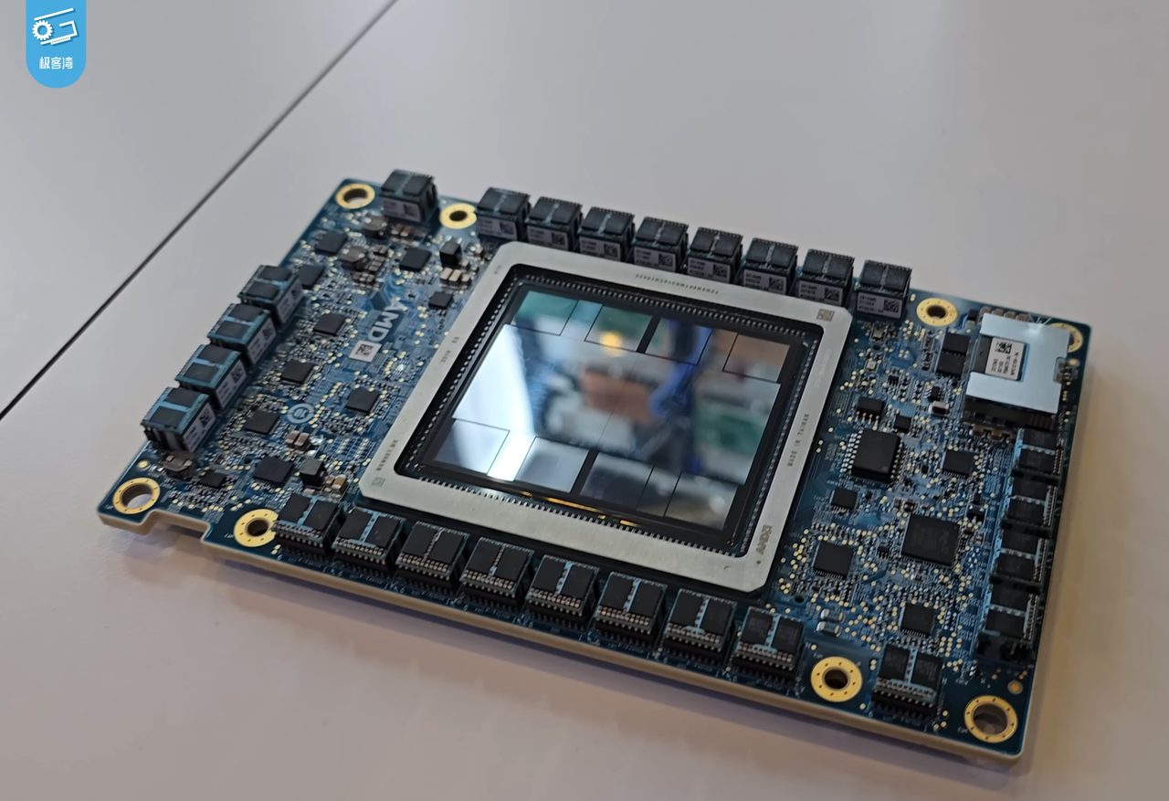 An AMD Instinct accelerator module lying on a white table: a blue circuit board with a large square package in a silver metal frame at its centre, the polished top of the package reflecting the room, rows of black components along the edges of the board, and an AMD logo printed on the board