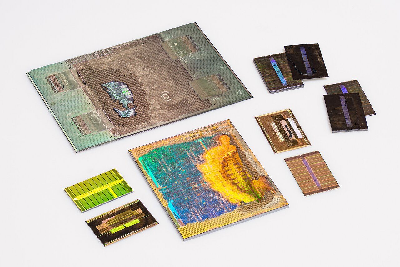 The parts of a Tesla P100 package laid out on a white surface: a large grey silicon slab, a rainbow-coloured GPU die, and several small rectangular memory dies with a stripe down the middle