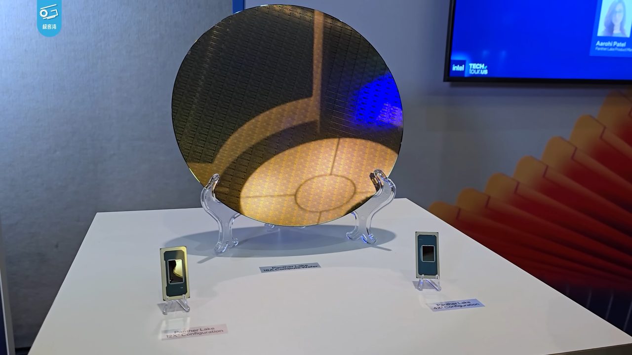 A silicon wafer standing upright in a clear stand on a white table at an exhibition, its surface reflecting orange and yellow light, with two small processor packages displayed on stands in front of it