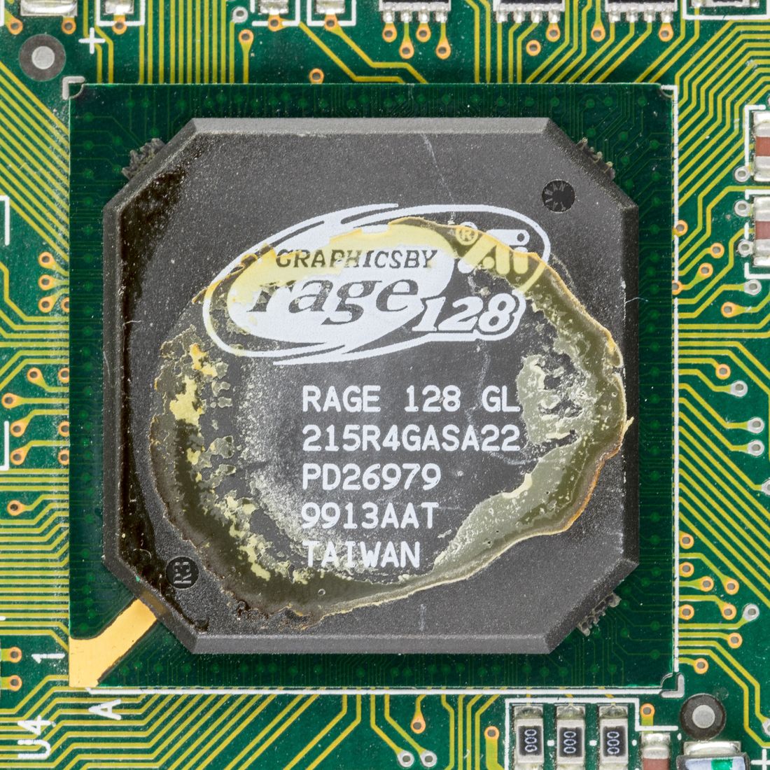 Close-up of an ATI Rage 128 GL graphics chip on a green circuit board, its black package printed with the Rage 128 logo and the words Graphics by ATI, partly covered by a dried yellowish layer of old thermal paste