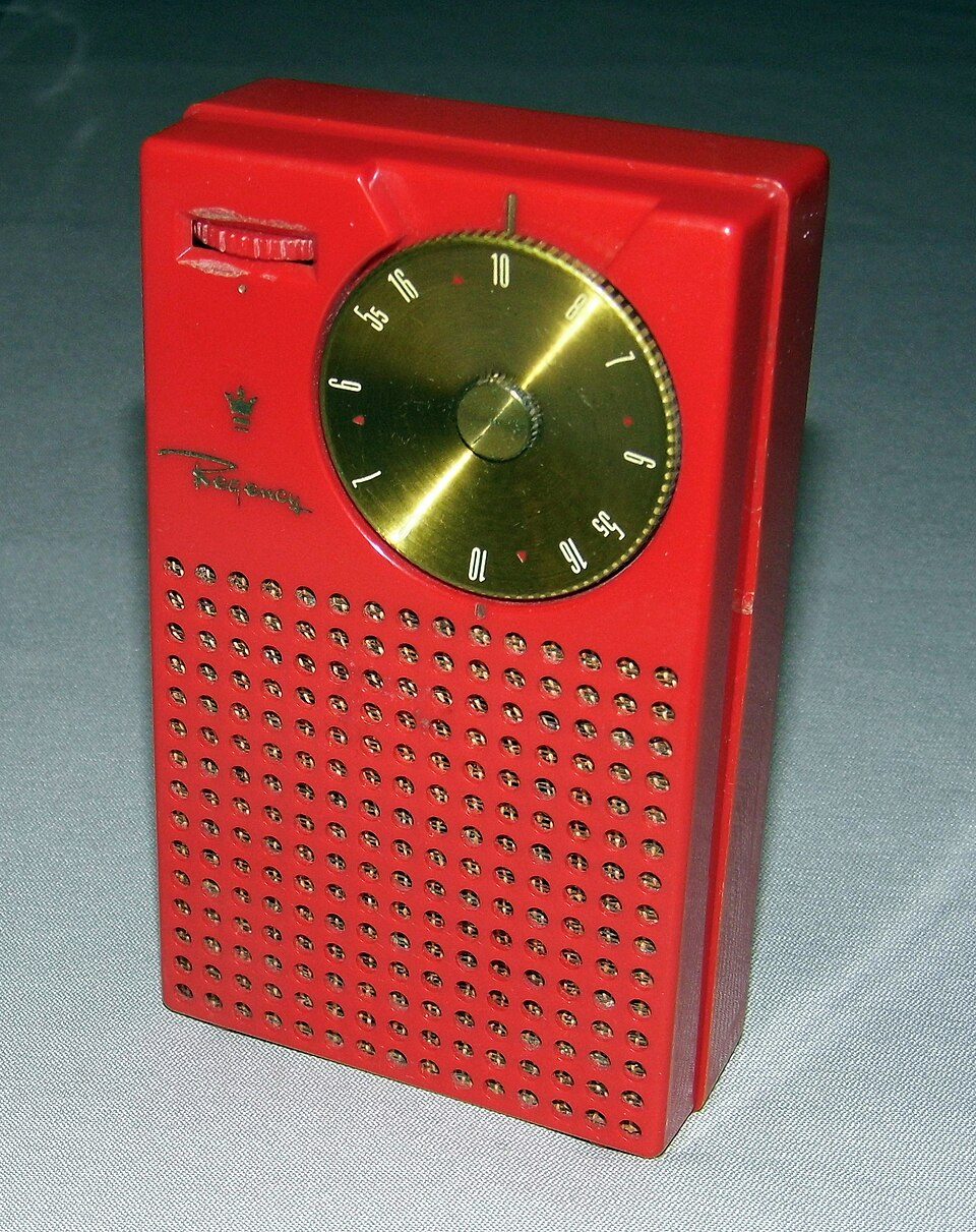 A red Regency TR-1 pocket radio: a small rectangular case with a gold tuning dial and a grid of round speaker holes