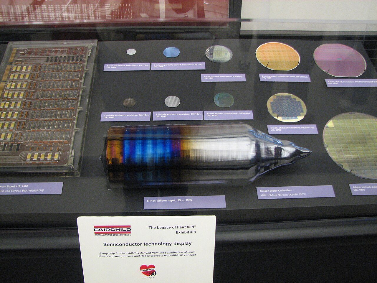 A museum display case with a polished silicon ingot lying in front and wafers of increasing diameter arranged behind it