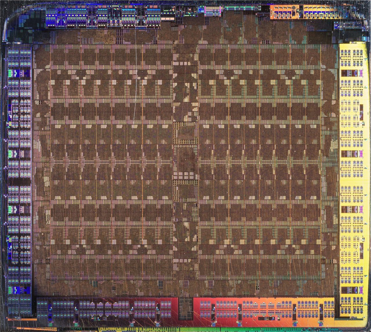 Photograph of the AMD Tahiti die: a large brown grid of repeated rectangular blocks in two halves filling the middle, with bands of differently coloured circuitry, blue, yellow, orange and red, along all four edges