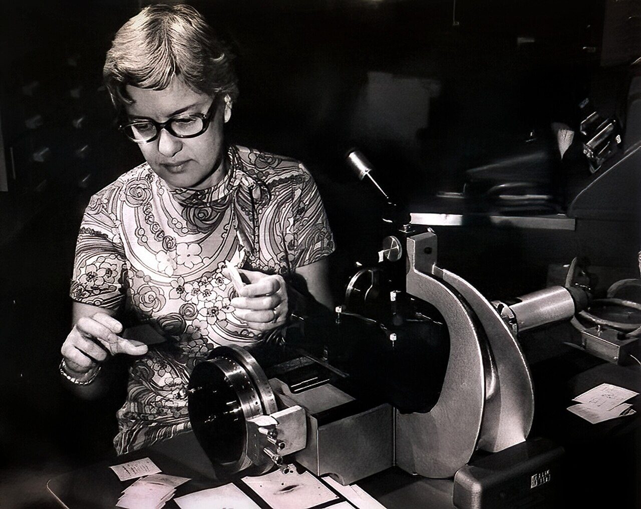Vera Rubin in 1974, wearing glasses, examining a glass plate of spectra at a measuring machine